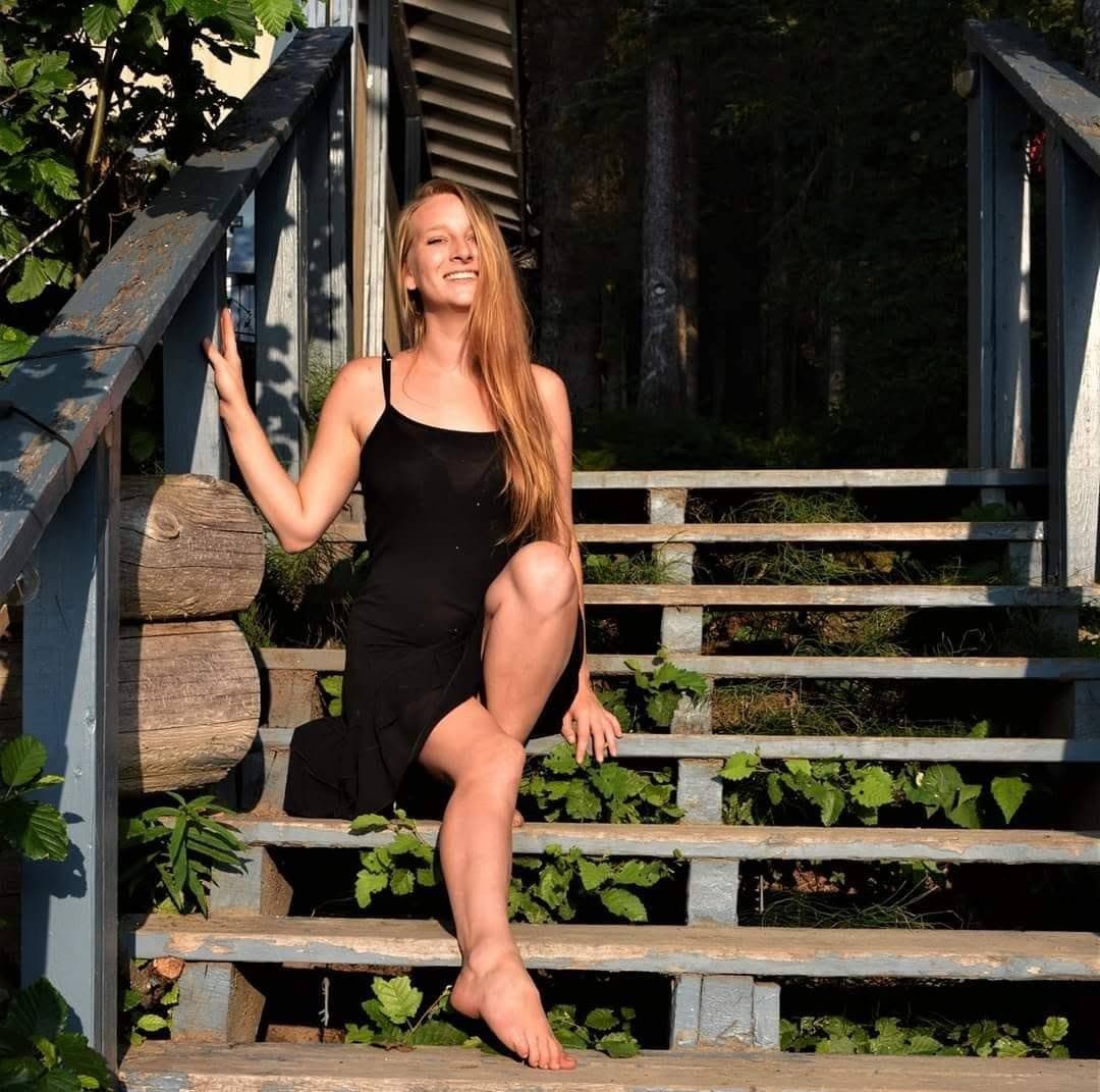 Smiling woman with long blonde hair sits on rustic wooden stairs wearing a black dress.