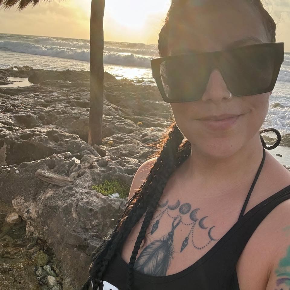 Woman with braided hair and chest tattoo wearing sunglasses on a rocky beach at sunset.